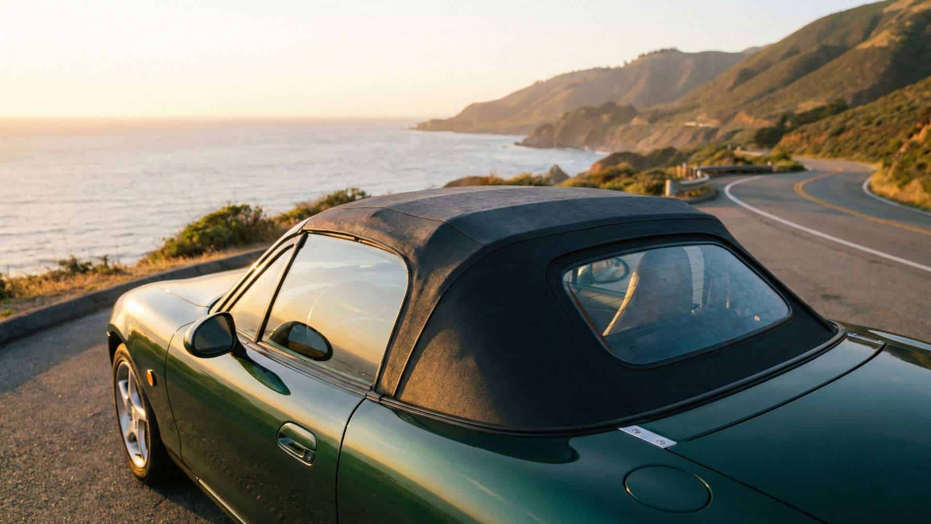 Mazda MX-5 NB verte, capote vinyle fermée, stationnée au bord d’une route côtière au coucher du soleil