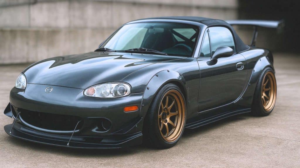 Mazda MX-5 NBFL tuning gris anthracite avec kit large, jantes bronze, splitter avant et aileron arrière, photographiée en 16/9 dans un décor urbain