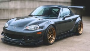 Mazda MX-5 NBFL tuning gris anthracite avec kit large, jantes bronze, splitter avant et aileron arrière, photographiée en 16/9 dans un décor urbain