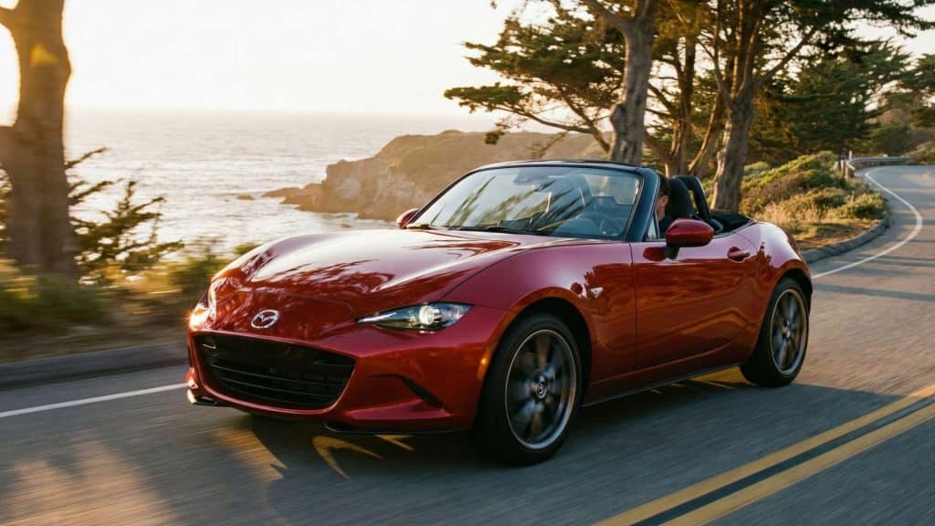 Mazda MX-5 ND rouge roulant au bord de la mer sur une route sinueuse au coucher du soleil