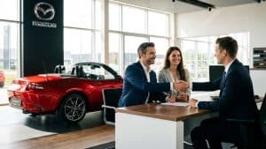 Signature d’un contrat de vente dans une concession Mazda avec un couple et un vendeur devant une Mazda MX-5 rouge décapotable.