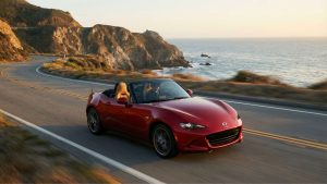 Cabriolet Mazda MX-5 rouge roulant sur une route côtière sinueuse au coucher du soleil avec l'océan et des falaises en arrière-plan.