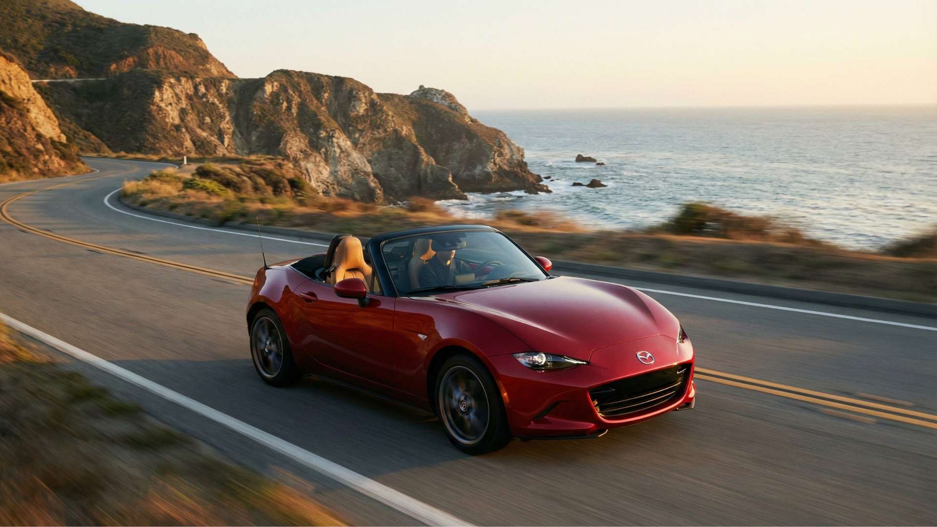Cabriolet Mazda MX-5 rouge roulant sur une route côtière sinueuse au coucher du soleil avec l'océan et des falaises en arrière-plan.