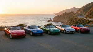 Cinq générations de Mazda MX-5 (NA, NB, NBFL, NC, ND) décapotées, garées côte à côte au coucher du soleil sur un parking surplombant une route côtière sinueuse et l'océan Pacifique