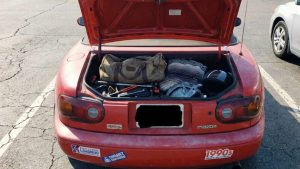 Coffre ouvert d'une Mazda MX-5 NA rouge, rempli de sacs de voyage, d'outils et d'un casque, pris en photo de manière amateur avec un smartphone dans un parking