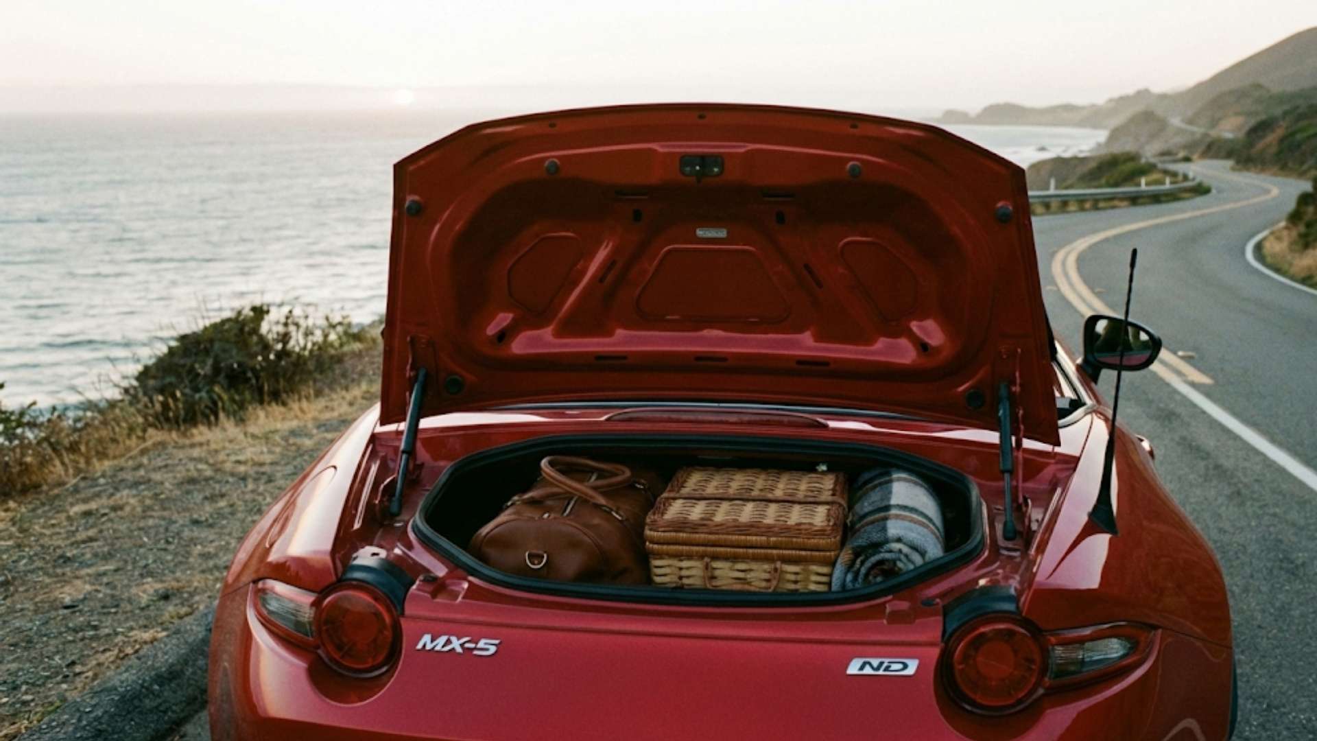 Coffre ouvert d'une Mazda MX-5 rouge garée au bord d'une route côtière, contenant un panier en osier et un sac en cuir, sous un ciel de coucher de soleil