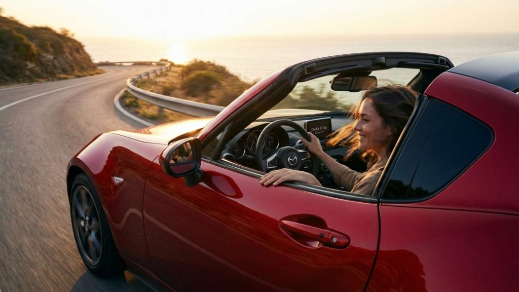 Femme souriante conduisant un cabriolet Mazda MX-5 rouge décapoté sur une route côtière sinueuse au coucher du soleil, incarnant le concept de Jinba Ittai (l'unité entre le cheval et le cavalier)