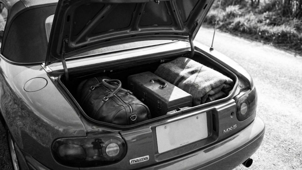 Le coffre ouvert d'une Mazda MX-5 NB (miata) est rempli d'un sac de voyage en cuir, d'une petite caisse et d'une couverture, le tout photographié en noir et blanc pour une ambiance classique
