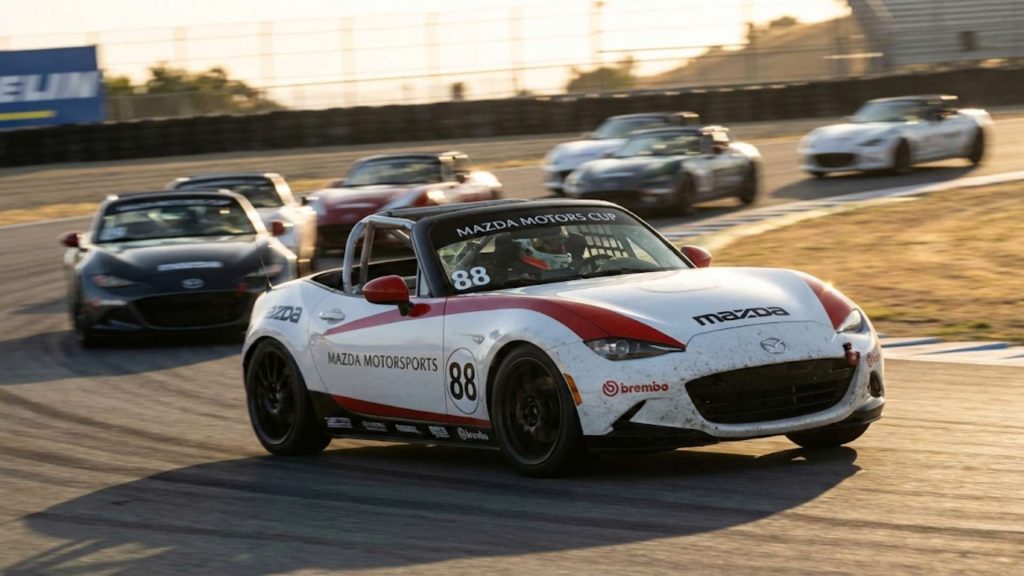 Gros plan sur une Mazda MX-5 Cup blanche et rouge de course, le numéro 88 bien visible, menant un peloton d'autres voitures de course MX-5 sur un circuit au coucher du soleil.