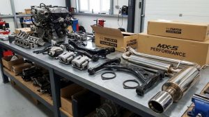 Vue d'ensemble d'un atelier spécialisé dans les pièces de Mazda MX-5, montrant plusieurs voitures en réparation, des mécaniciens travaillant sur les moteurs, et des étagères remplies de pièces de rechange, y compris des moteurs, des systèmes d'échappement et des suspensions