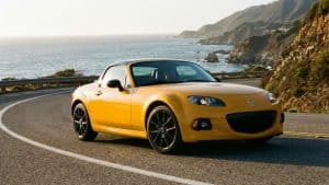 Une Mazda MX-5 NC jaune décapotable roulant sur une route sinueuse en bord de mer avec des falaises en arrière-plan.
