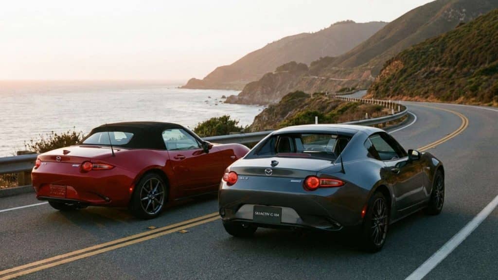 Deux Mazda MX-5, une rouge et une grise RF, stationnées sur une route côtière sinueuse au coucher du soleil face à l’océan.