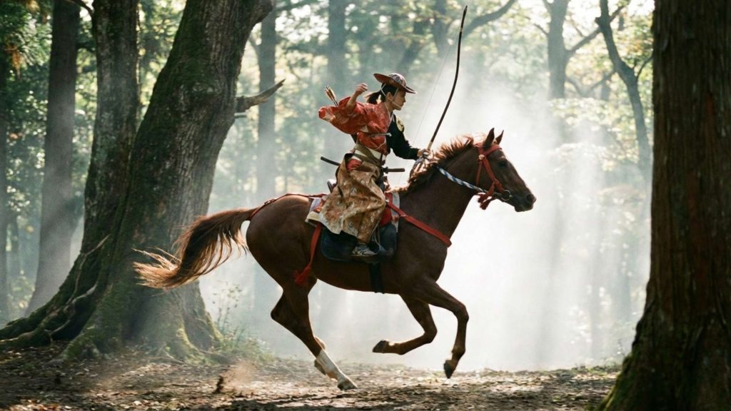 Un cavalier japonais traditionnel, vêtu d'une tenue de chasse à cheval (yabusame), galope à travers une forêt brumeuse, tenant un arc et des flèches, illustrant le concept de Jinba Ittai, l'unité du cheval et du cavalier