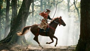 Un cavalier japonais traditionnel, vêtu d'une tenue de chasse à cheval (yabusame), galope à travers une forêt brumeuse, tenant un arc et des flèches, illustrant le concept de Jinba Ittai, l'unité du cheval et du cavalier