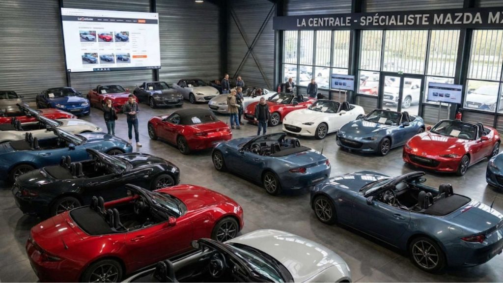 Un grand garage spécialisé rempli de nombreux modèles de Mazda MX-5 de différentes couleurs et générations, avec des personnes qui les regardent.