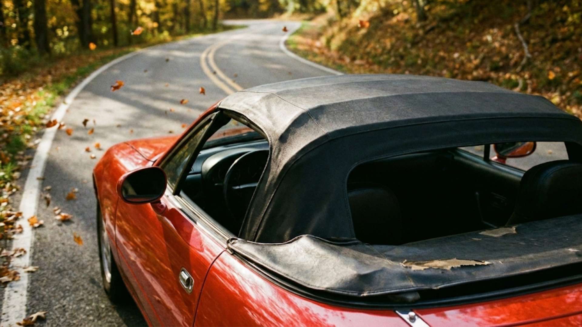 Une image photoréaliste au format 16/9 d'une Mazda MX-5 rouge avec la capote noire relevée, roulant sur une route sinueuse bordée d'arbres aux couleurs automnales, avec des feuilles mortes tombant sur le capot.