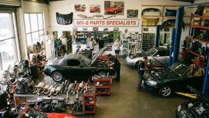 Vue d'ensemble d'un atelier spécialisé dans les pièces de Mazda MX-5, montrant plusieurs voitures en réparation, des mécaniciens travaillant sur les moteurs, et des étagères remplies de pièces de rechange, y compris des moteurs, des systèmes d'échappement et des suspensions