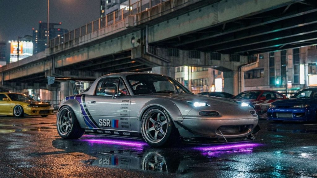 Mazda MX-5 NA grise préparée avec kit widebody, jantes sport et néons violets, photographiée de nuit sous un pont en environnement urbain