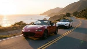 Une Mazda MX-5 rouge et une Fiat 124 Spider blanche roulant côte à côte sur une route côtière sinueuse baignée par la lumière dorée du crépuscule.