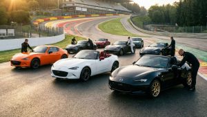 Une collection de Mazda MX-5 en éditions spéciales, incluant la 30ème Anniversaire orange et la Eunos Edition noire, alignées avec leurs pilotes sur le circuit de Spa-Francorchamps au coucher du soleil