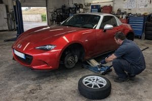 Photo réaliste d’une Mazda MX-5 ND rouge à toit argent dans un garage pro, avec un mécano en train de changer une roue, prise façon smartphone en lumière naturelle