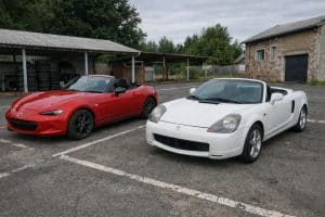 Mazda MX-5 rouge garée à côté d’une Toyota MR2 blanche sur un parking extérieur pour un comparatif entre deux roadsters japonais