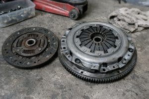 Photo réaliste d’un embrayage Mazda MX-5 démonté avec disque, mécanisme et volant moteur posés sur le sol d’un atelier, prise en style Panasonic GH4 avec lumière naturelle et rendu documentaire