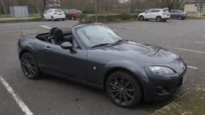 Mazda MX-5 NC grise garée sur une place de parking, capote ouverte, vue avant latérale, dans un environnement urbain sobre sous ciel nuageux