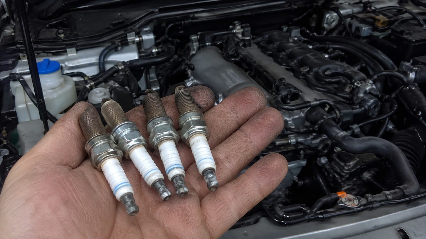 Main tenant quatre bougies d’allumage démontées devant le compartiment moteur d’une Mazda MX-5 ND ouverte, photo réaliste de maintenance automobile prise en lumière naturelle.