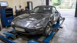 Mazda MX-5 NA gris foncé photographiée dans un atelier automobile pendant un contrôle de géométrie et de parallélisme, installée sur un pont de réglage avec têtes de mesure sur les roues, lumière naturelle et rendu documentaire réaliste.
