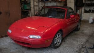 Cote et investissement Mazda MX-5 : le guide complet 2026 4 Mazda MX-5 NA rouge stationnée dans un garage, vue avant trois-quarts, avec capote noire, phares escamotables fermés, sol en béton et atelier de rangement en arrière-plan