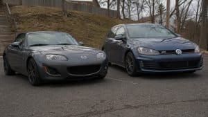 Mazda MX-5 NC gris foncé garée à côté d’une Volkswagen Golf GTI bleu foncé sur un parking extérieur, comparaison visuelle entre roadster sportif propulsion et compacte sportive GTI sous lumière naturelle.