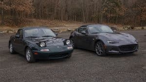 Mazda MX-5 NA avec phares escamotables à côté d’une Mazda MX-5 ND grise moderne, comparatif visuel entre deux générations du roadster japonais sur parking boisé.
