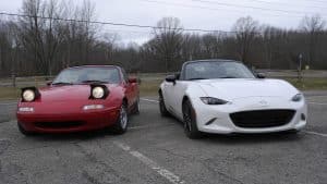 Mazda MX-5 NA rouge avec phares escamotables relevés garée à côté d’une Mazda MX-5 ND blanche moderne sur un parking extérieur, comparaison visuelle entre première et quatrième génération du roadster japonais sous ciel couvert.