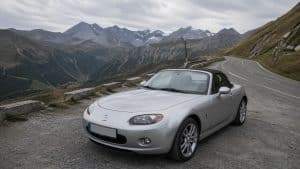 Mazda MX-5 NC grise garée sur un col de montagne, avec route alpine sinueuse, virages en lacets, barrières de sécurité et sommets rocheux en arrière-plan.