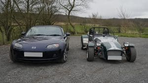 Photo Panasonic GH4 montrant une Mazda MX-5 NC bleu foncé garée à côté d’une Caterham Seven verte et argent sur un parking gravillonné, comparaison visuelle entre deux roadsters légers sportifs sous ciel couvert.