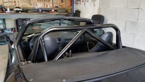 Arceau Mazda MX5 : sécurité, prix et choix par génération 3 Arceau de sécurité homologué monté sur une Mazda MX-5 NBFL noire dans un atelier mécanique. La photo montre l’intérieur du roadster décapotable avec un arceau tubulaire en acier noir reliant les points de fixation arrière derrière les sièges. Équipement de sécurité utilisé pour la conduite sportive, le circuit, le drift et les trackdays afin d'améliorer la protection contre le retournement du véhicule.
