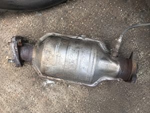 Catalyseur Mazda MX-5 : symptômes, coût et vol expliqués 2 Catalyseur Mazda MX-5 NBFL 1.8L essence démonté lors d'une réparation mécanique, posé sur un sol d'atelier, montrant le corps du catalyseur, la sonde lambda, les brides de fixation et des traces de corrosion superficielles sur le système d'échappement d'origine Mazda.