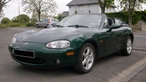 Photo d'une Mazda MX-5 NBFL verte British Racing Green, roadster cabriolet deux places produit par Mazda entre 2001 et 2005, vue de trois quarts avant dans un environnement résidentiel. On distingue les phares ovales caractéristiques du restylage NBFL, les jantes aluminium d'origine, la capote ouverte et la carrosserie verte brillante. La voiture est stationnée sur une route de quartier avec plusieurs véhicules et maisons en arrière-plan, dans une ambiance documentaire réaliste.