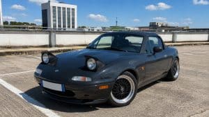 Photo format 16:9 d’une Mazda MX-5 NA noire en plein soleil sur un parking rooftop urbain, avec phares escamotables ouverts, jantes type BBS noires à bord poli, carrosserie brillante, capote noire et ombres marquées au sol, illustration visuelle du bruit Mazda MX-5 et d’un roadster japonais youngtimer en extérieur.