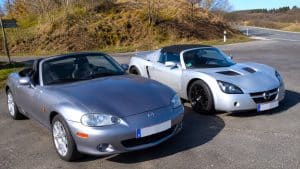 Mazda MX-5 vs Opel Speedster : le duel roadster qui surprend 1 Mazda MX-5 NB décapotable grise et Opel Speedster argentée stationnées côte à côte sur une aire routière ensoleillée, comparaison du gabarit et du design des deux roadsters sportifs, photographie amateur Panasonic GH4, 15 juillet 2026, format 16:9