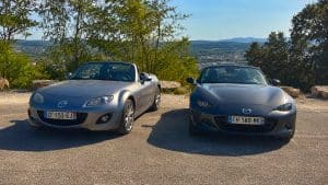 MX-5 NC vs ND : quelle génération choisir vraiment 2 Comparatif Mazda MX-5 NC grise à gauche et Mazda MX-5 ND gris foncé à droite, deux cabriolets décapotés photographiés de face sur un parking panoramique ensoleillé avec arbres, ville et collines en arrière-plan, 17 juillet 2026, format 16:9