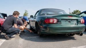 Deux hommes accroupis examinant le moyeu arrière sans roue d'une Mazda MX-5 NB verte à hardtop en rassemblement
