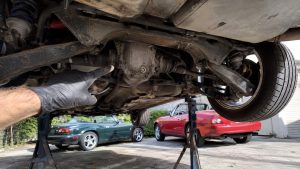 Inspection du différentiel arrière d’une Mazda MX-5 sur chandelles, main gantée pointant le carter de transmission sous le châssis, bras de suspension, cardans et roue arrière visibles, deux Mazda MX-5 de générations différentes stationnées en arrière-plan sous un soleil estival, photographie automobile documentaire réaliste, 2026, format 16:9