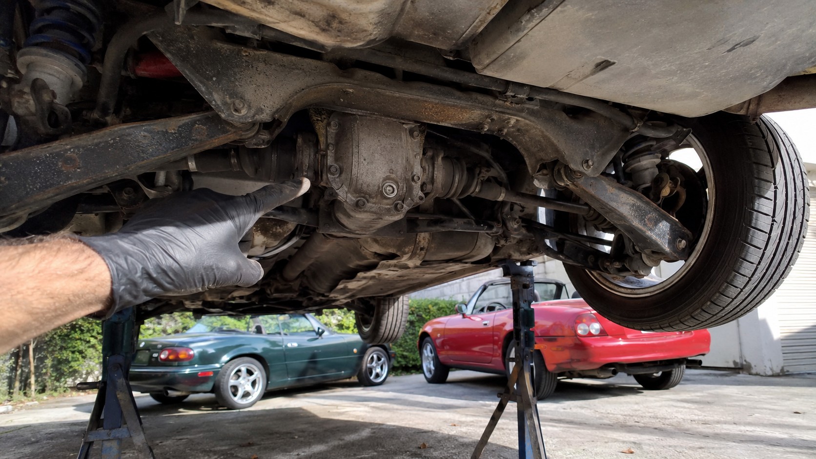Inspection du différentiel arrière d’une Mazda MX-5 sur chandelles, main gantée pointant le carter de transmission sous le châssis, bras de suspension, cardans et roue arrière visibles, deux Mazda MX-5 de générations différentes stationnées en arrière-plan sous un soleil estival, photographie automobile documentaire réaliste, 2026, format 16:9