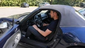 Mazda MX-5 grande taille : allez-vous vraiment rentrer dedans 2 Mazda MX-5 ND bleue avec capote souple, conducteur de grande taille installé dans l’habitacle compact, jambes repliées et espace intérieur limité, portière ouverte sur un parking ensoleillé, photo Canon EOS R6, 2026, format 16:9