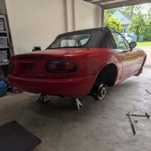 Photographie carrée de 1254 x 1254 px prise dans un garage domestique. Mazda MX-5 de première génération NA, rouge vif, capote souple noire fermée et délavée, vue de trois quarts arrière. Le train arrière repose sur deux chandelles métalliques placées sous les points de levage, une roue arrière déposée : moyeu, disque de frein piqué de rouille et étrier apparents. Hayon avec logo MAZDA embossé, emplacement de plaque vide, feux arrière rectangulaires à blocs rouge, orange et blanc, pare-chocs rouge marqué de rayures, rétroviseur extérieur noir. Local à mur clair, étagère métallique garnie de bacs plastique transparents, bacs de rangement verts et bleus, porte de garage relevée ouvrant sur un jardin arboré, une maison blanche, une voiture claire et des conteneurs verts. Au premier plan à droite, clé en croix chromée sur la dalle béton, tapis de sol noir dans l'angle inférieur gauche.