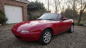 Mazda MX-5 NA rouge capote ouverte sur une allée de graviers devant un garage en briques