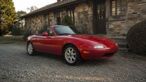 Mazda MX-5 NA rouge à jantes style Minilite devant une maison en pierre au soleil couchant