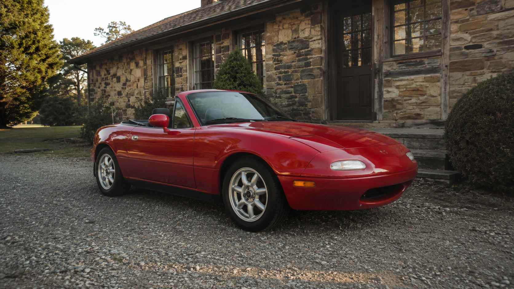 Mazda MX-5 NA rouge a jantes style Minilite devant une maison en pierre au soleil couchant